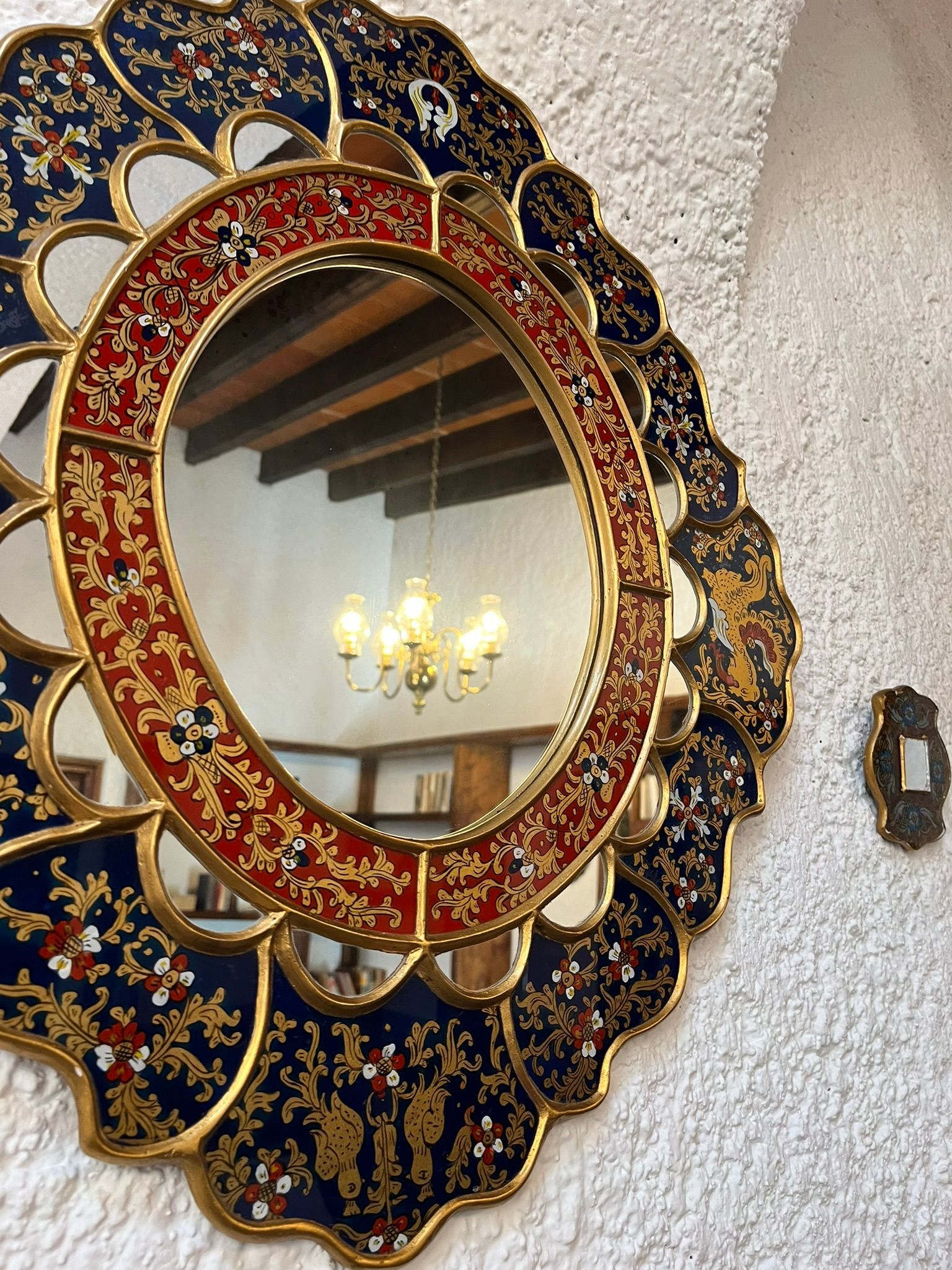 Un espejo ornamentado con detalles dorados y un borde estampado en azul y rojo cuelga de una pared blanca.
