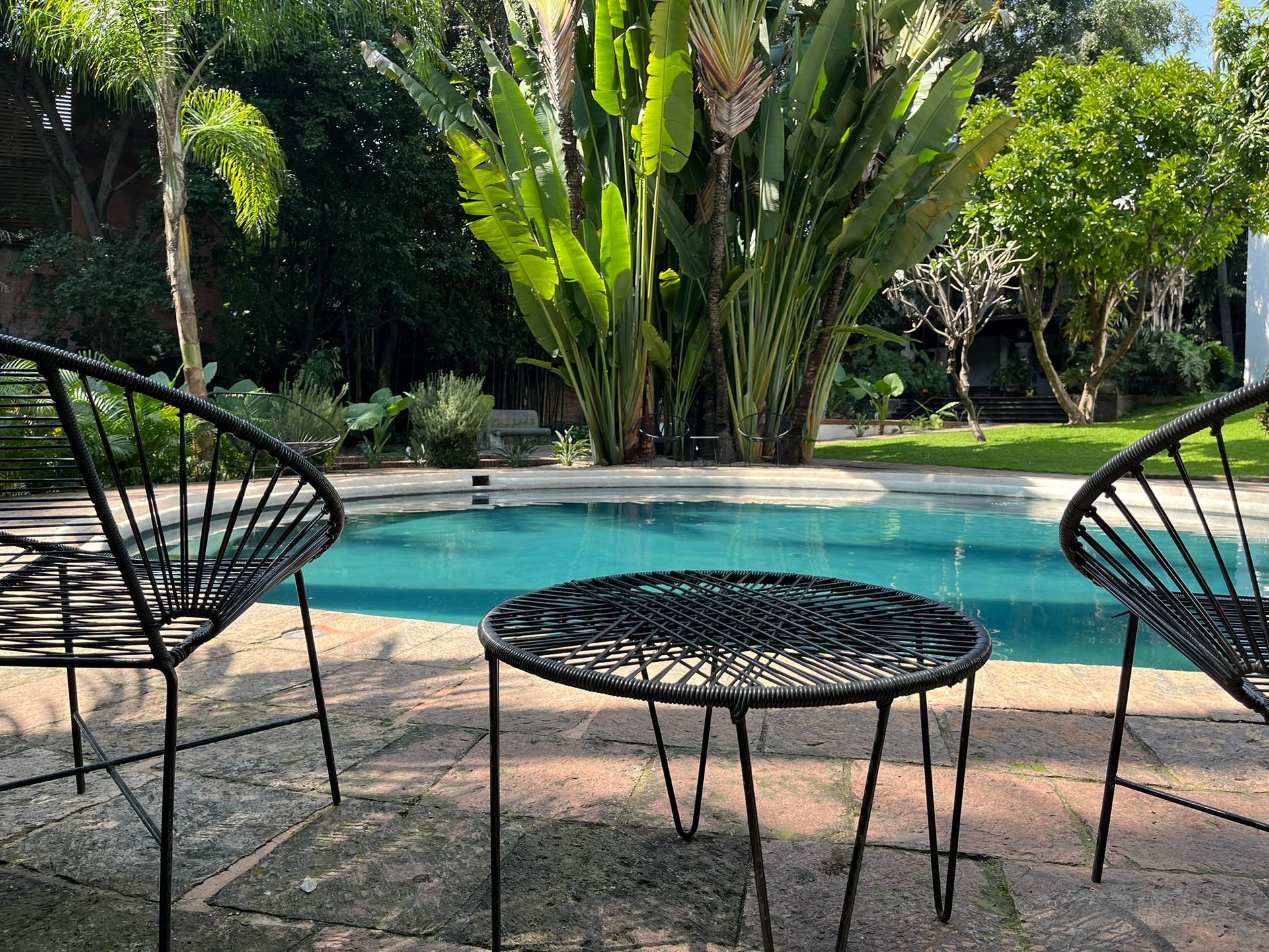 Dos sillas negras y una mesa junto a una piscina, plantas tropicales al fondo.
