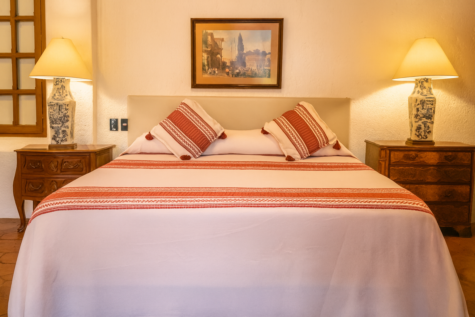 Dormitorio con cama, mesitas de noche, lámparas y un cuadro en la pared. La cama tiene sábanas rojas y blancas.