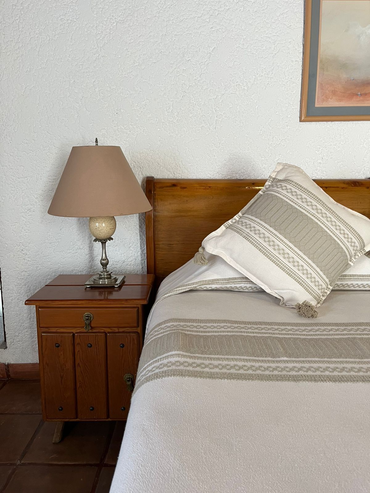 Dormitorio con mesita de noche de madera, cama, lámpara y pared blanca texturizada.