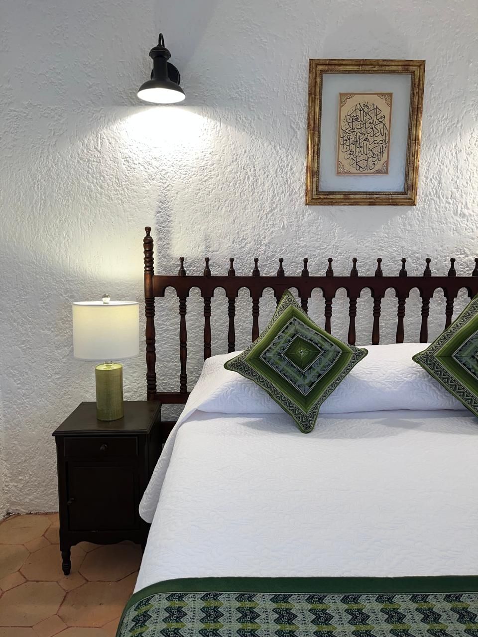 Dormitorio con cama blanca, cabecero de madera oscura, almohadas verdes y una pequeña mesita de noche con lámpara.