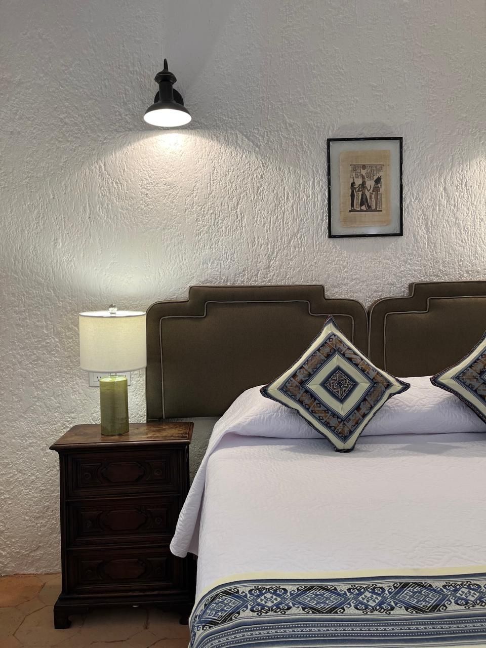 Dormitorio con pared blanca texturizada, cama, mesita de noche, lámparas y obras de arte.