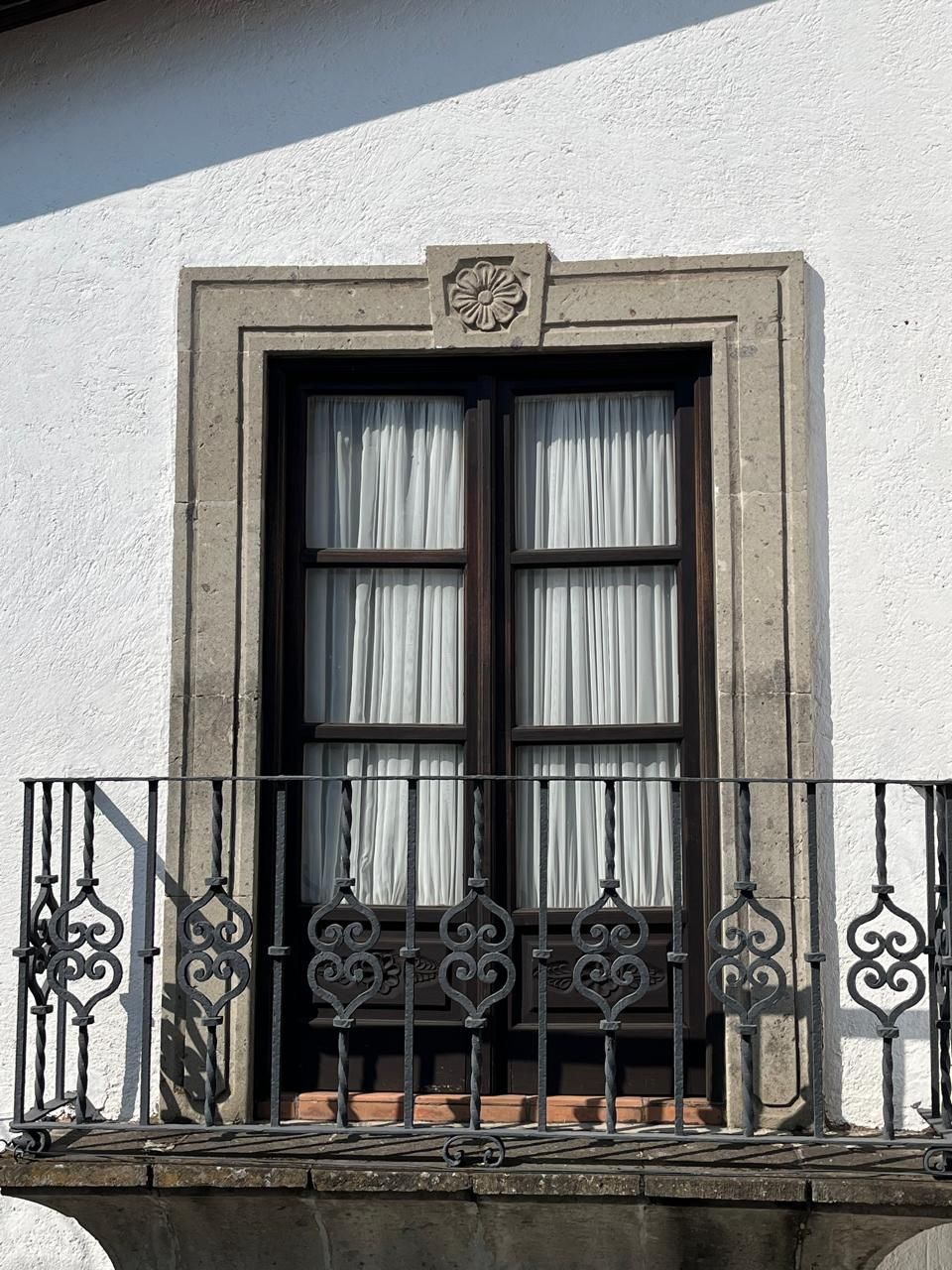 Ventana con marco de piedra decorativa, pequeño balcón con barandilla de hierro y cortinas blancas.