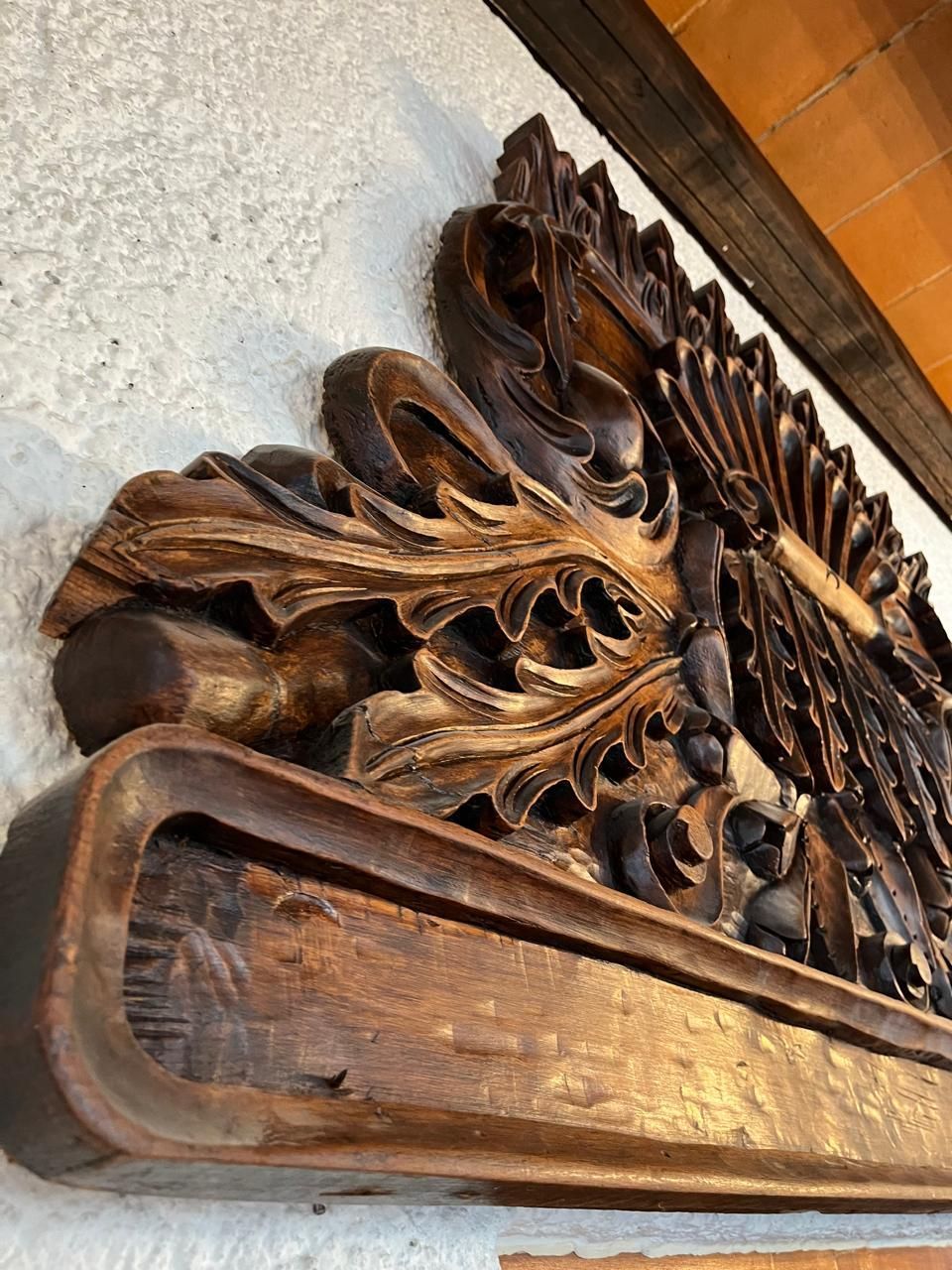 Detalle arquitectónico tallado en madera adornado montado sobre una pared blanca, con hojas estilizadas y elementos florales.