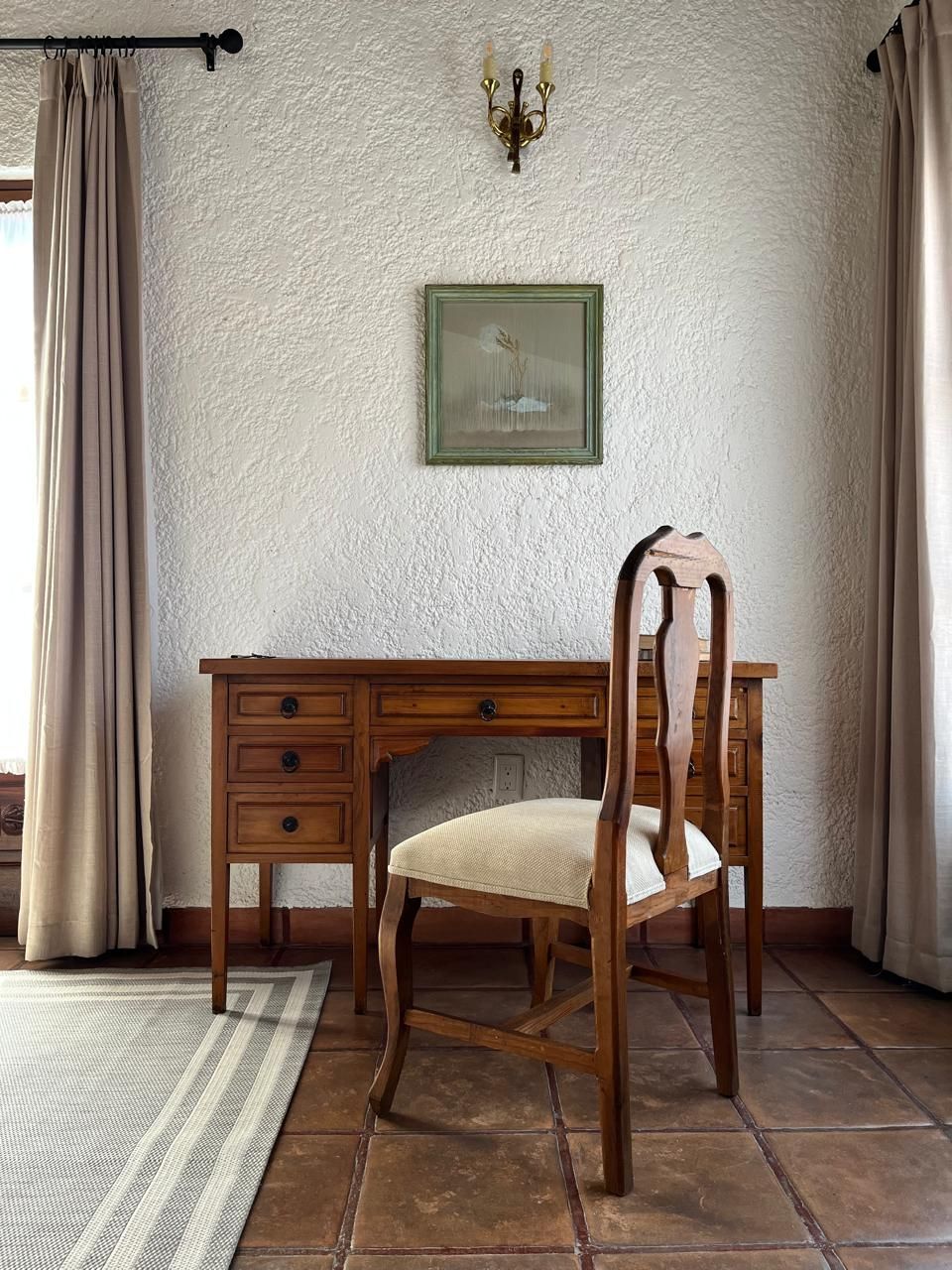Escritorio y silla de madera frente a una pared texturizada con un pequeño cuadro enmarcado y un aplique.