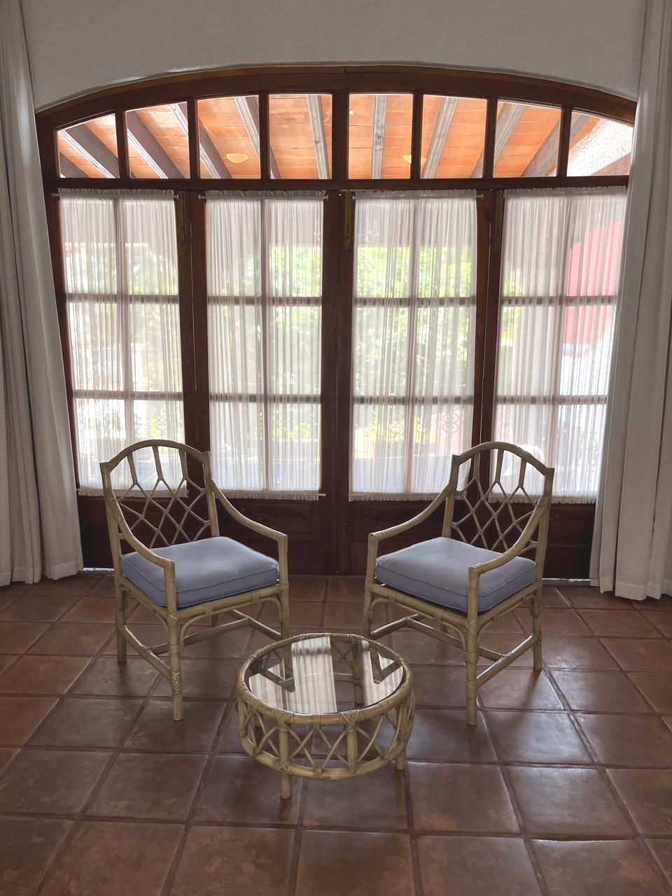 Dos sillas de mimbre y una mesa redonda frente a una gran ventana con cortinas transparentes.