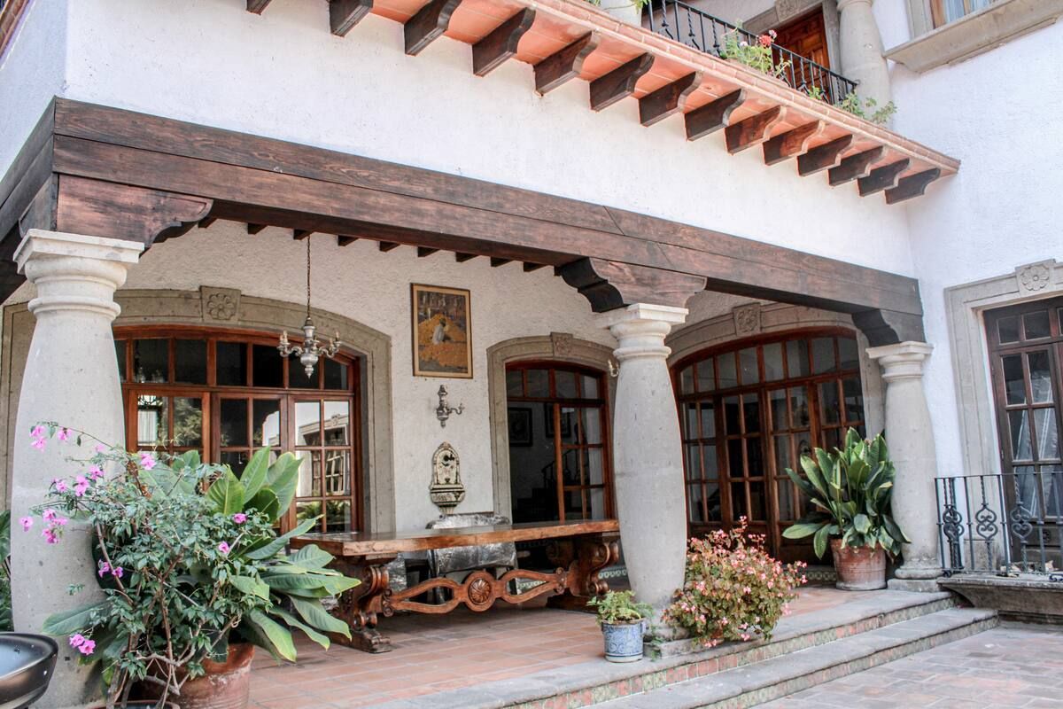 Patio cubierto con aberturas arqueadas y columnas de piedra, vigas de madera, plantas y una larga mesa de madera.