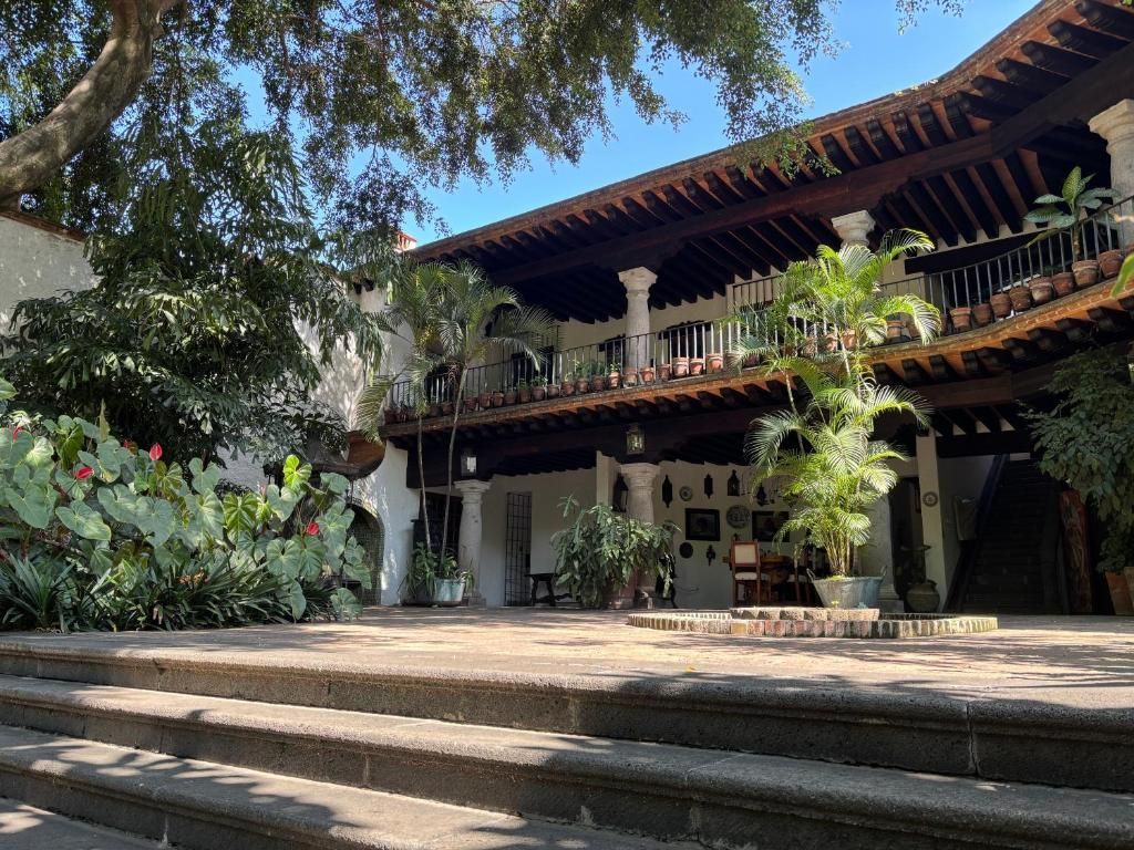 Edificio blanco de dos pisos con balcones de madera, exuberante vegetación y escalones de piedra bajo un cielo azul.