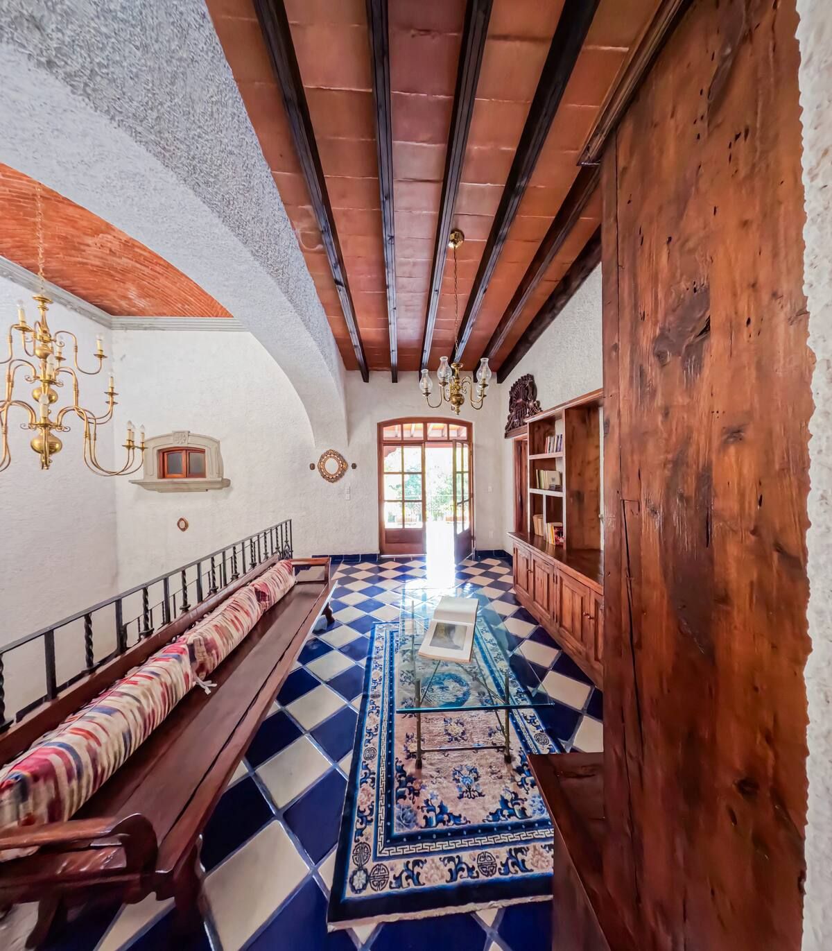 Pasillo largo con suelo de baldosas azules y blancas, vigas de madera, banco y candelabros antiguos.