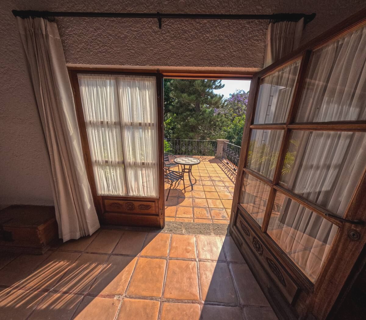 Puertas de madera abiertas que conducen a una terraza con mesa y sillas; la luz del sol entra a raudales en la habitación.