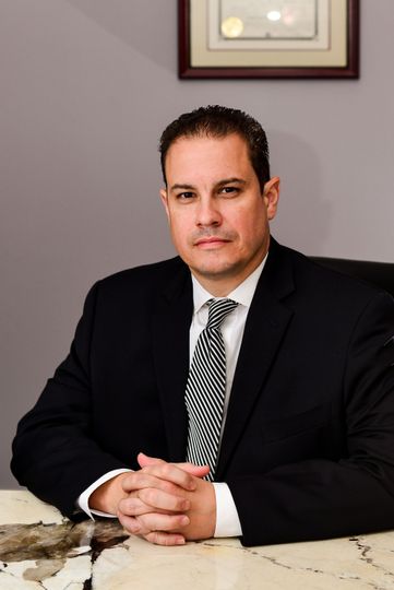 Man in a black suit seated at a marble table, hands clasped, in an office setting.