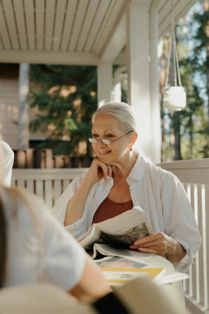 Smiling woman reading a book on a porch with another person seated nearby