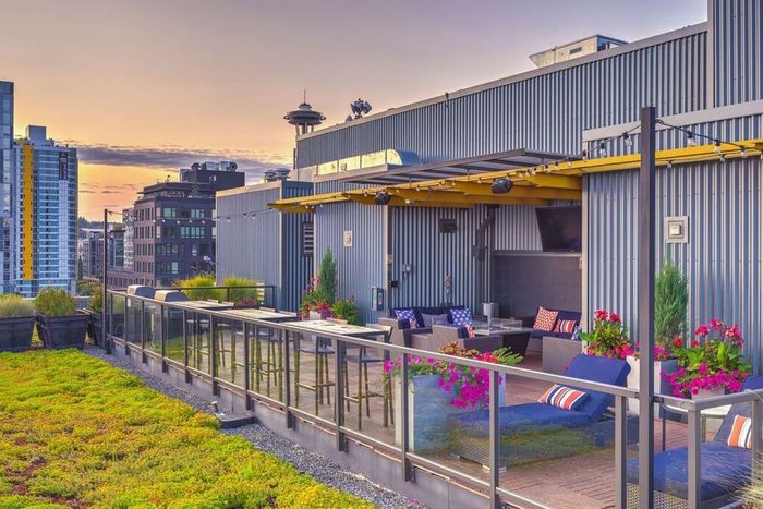 A rooftop lounge with outdoor seating, potted flowers, and a grassy terrace at sunset, featuring the Space Needle skyline.
