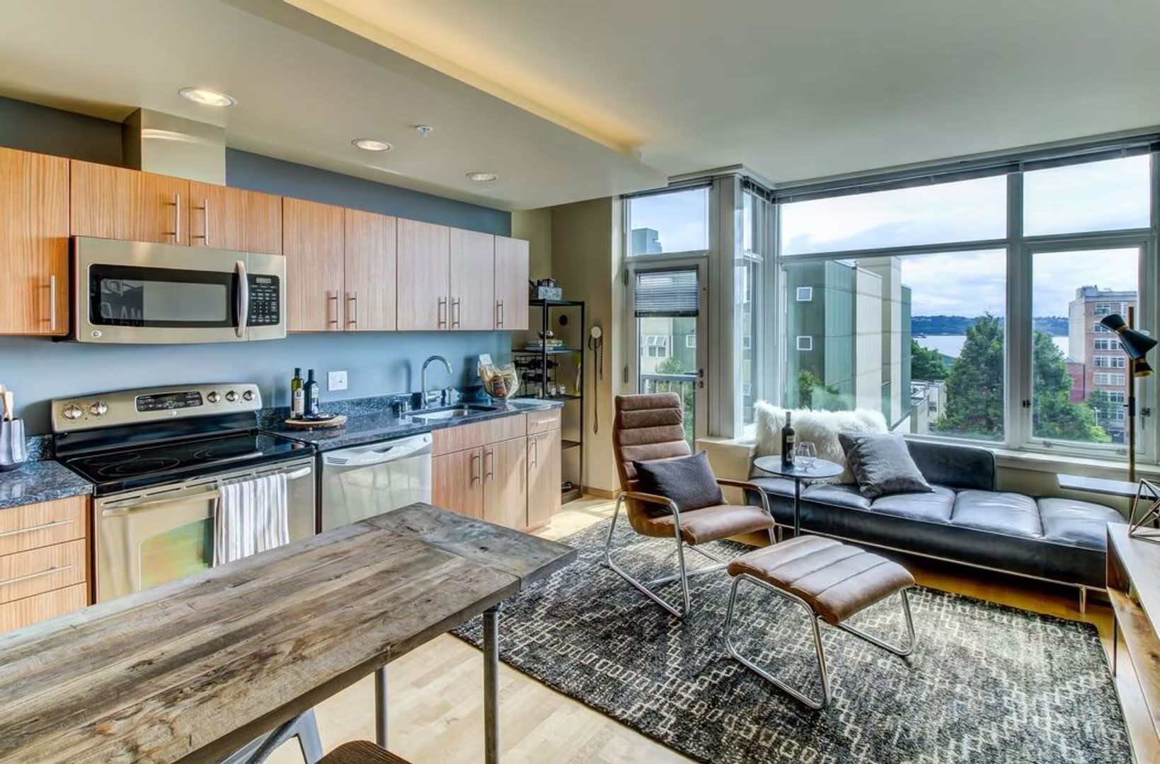 A modern kitchen with light wood cabinets and stainless steel appliances flows into a living area with a leather chair.