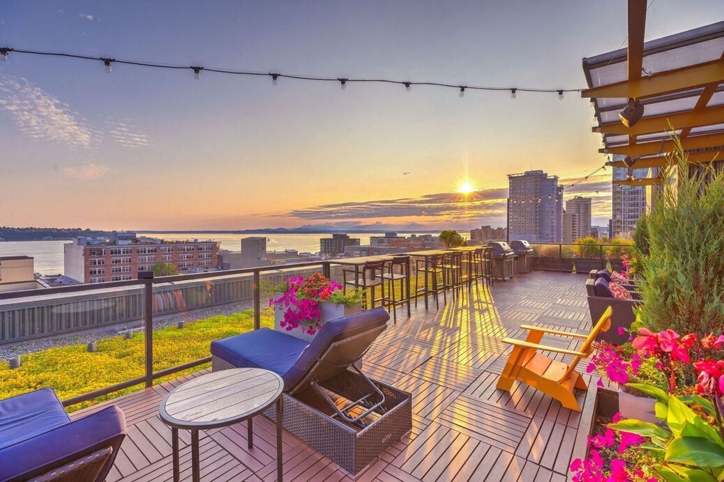 Rooftop outdoor lounge with seating, string lights, planters, and a city skyline at sunset.