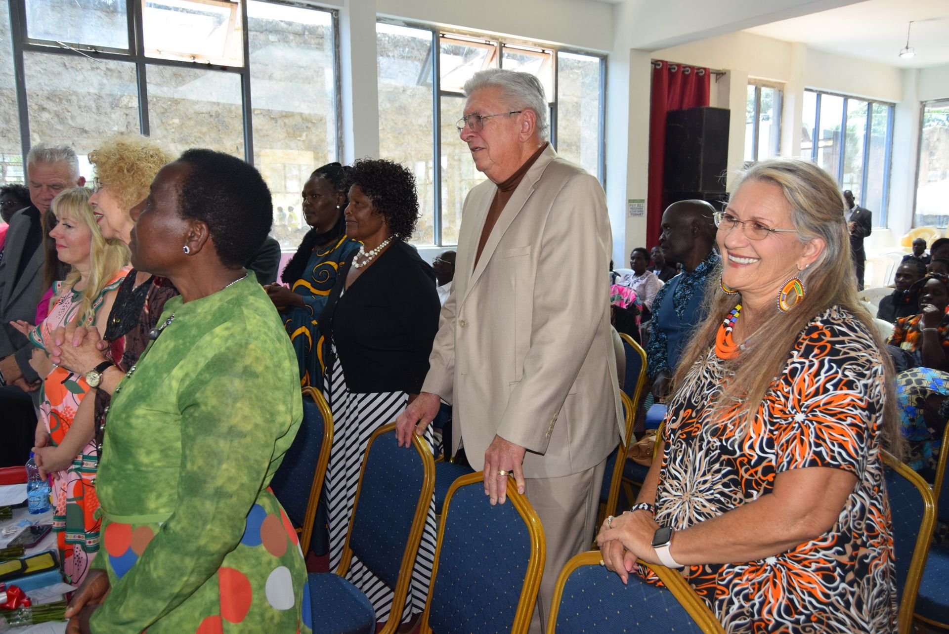 People standing, some smiling, in a bright room. A man in a tan suit is prominent. Others are clapping or looking on.