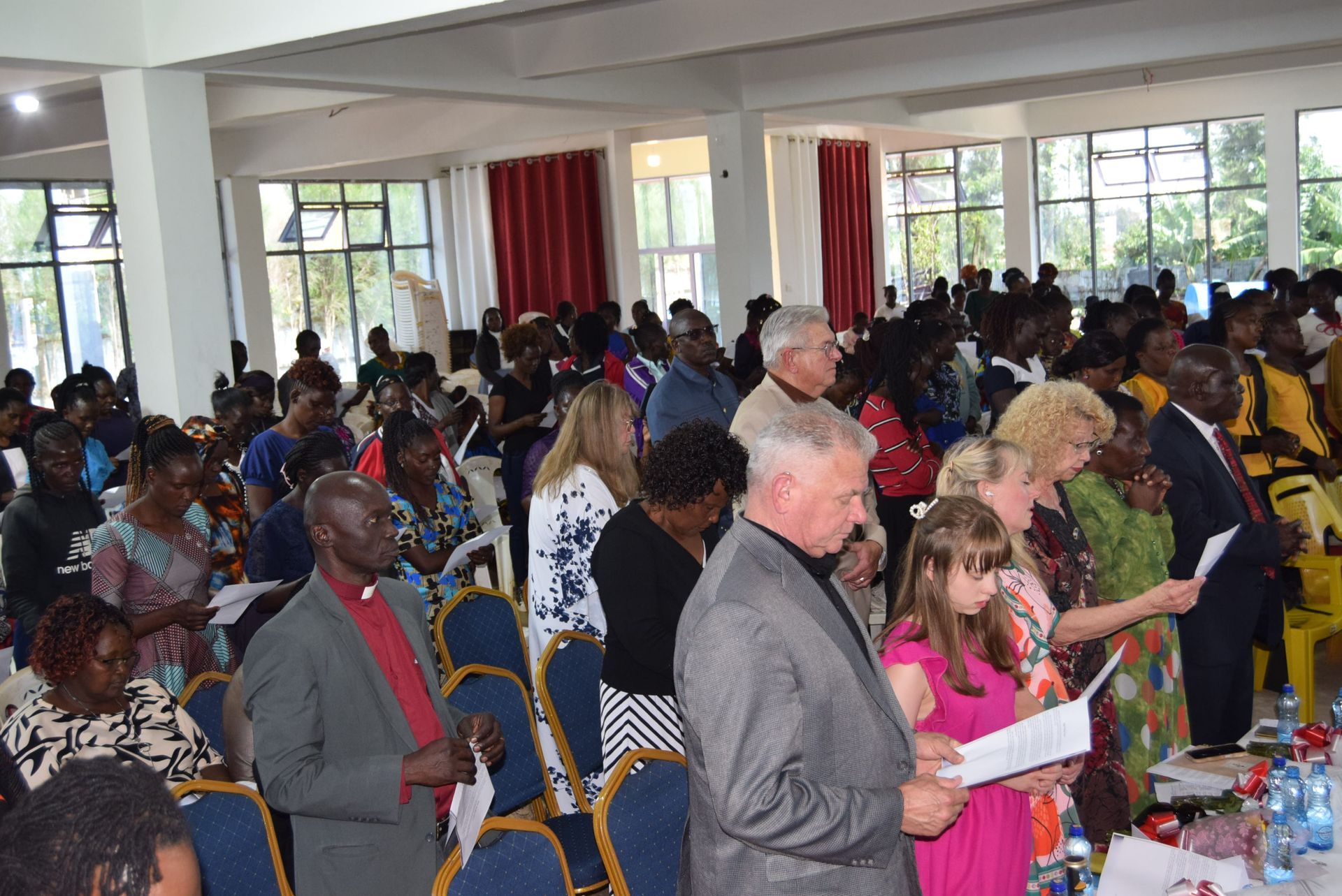 People standing and holding papers in a bright room, likely at a church service or conference.