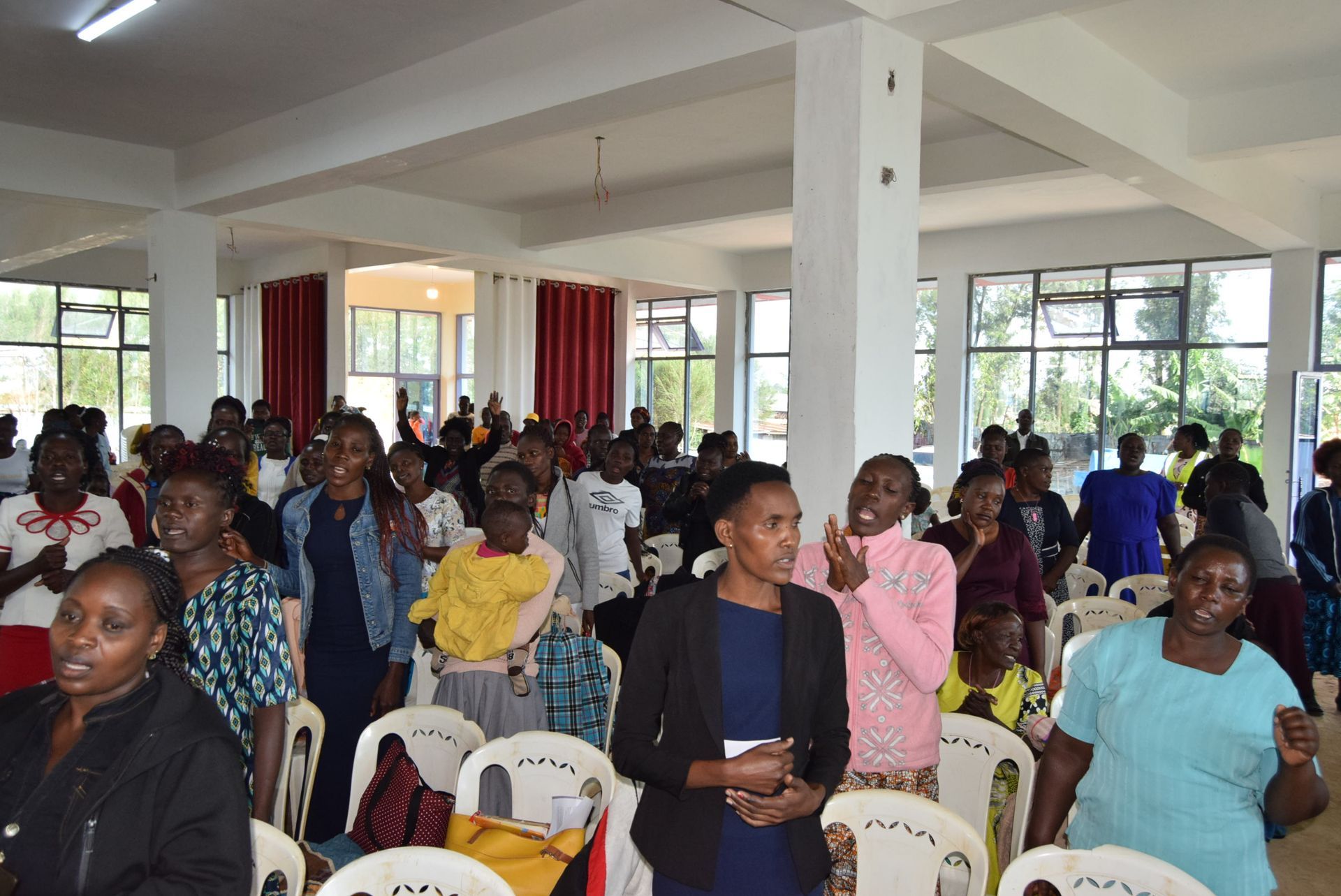 People in a bright hall, some singing, arms raised. Many seated in rows of white chairs.