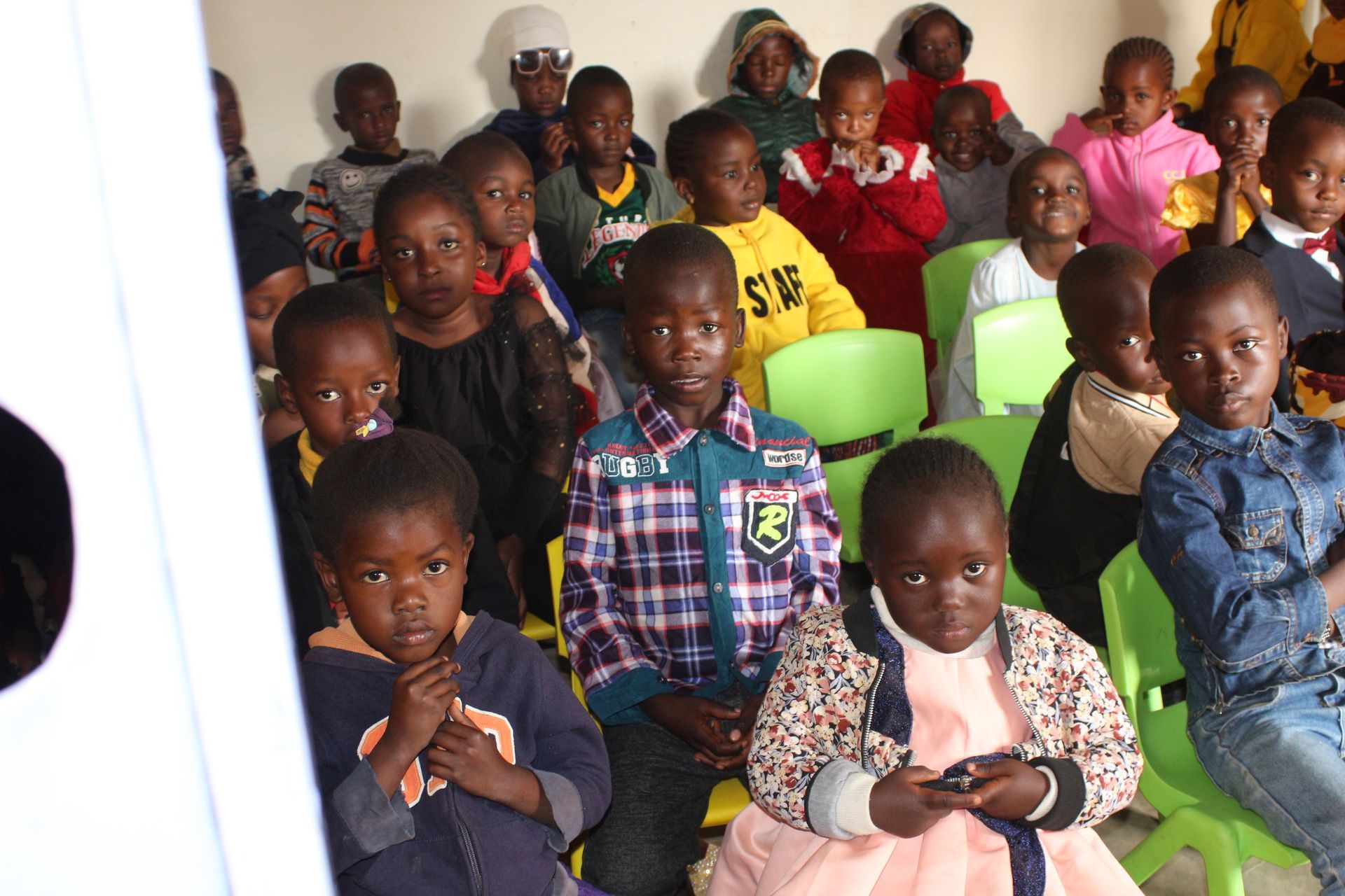 Children seated in a room, some wearing colorful clothes, looking at the camera. Green chairs and light walls.