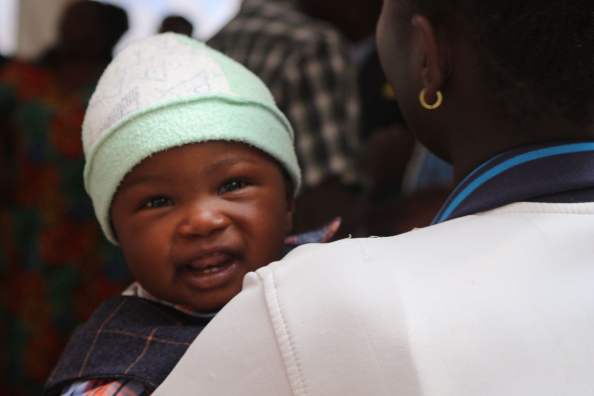 Baby smiling, wearing a green and white hat, held by someone in a white shirt, outdoors.