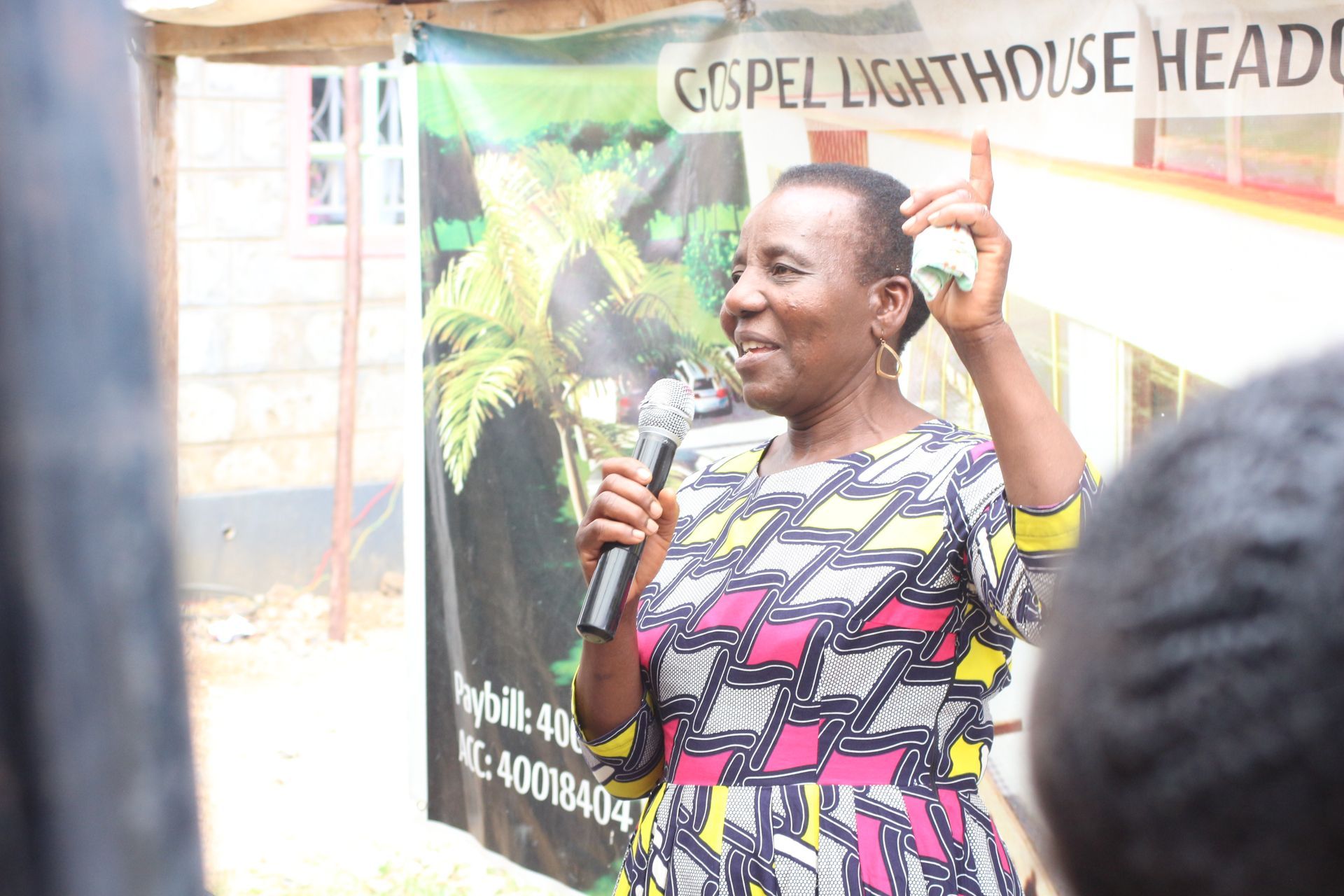 Woman speaking into a microphone outdoors, raising one hand. Gospel Lighthouse banner visible behind her.