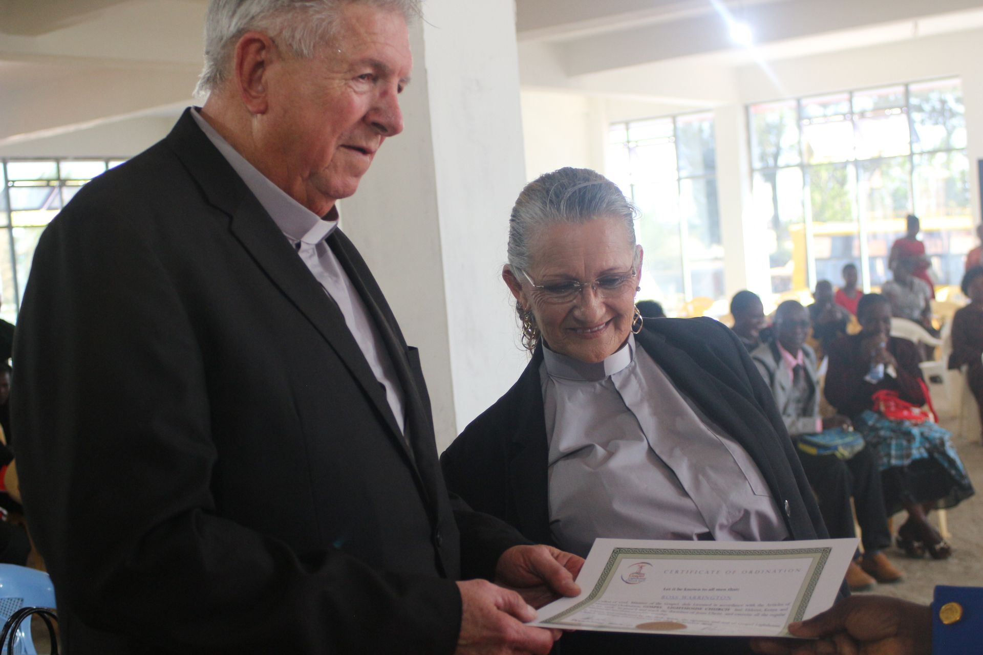 Two people examining a document. Man in a black suit. Woman in glasses wearing a grey shirt. Background: event with people.