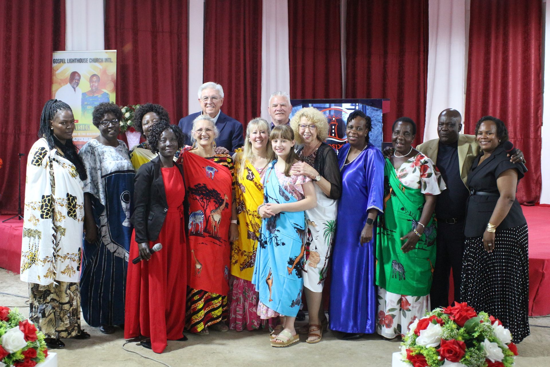 Group of people, mostly women, posing for a photo indoors, some wearing colorful dresses, red curtains in the background.