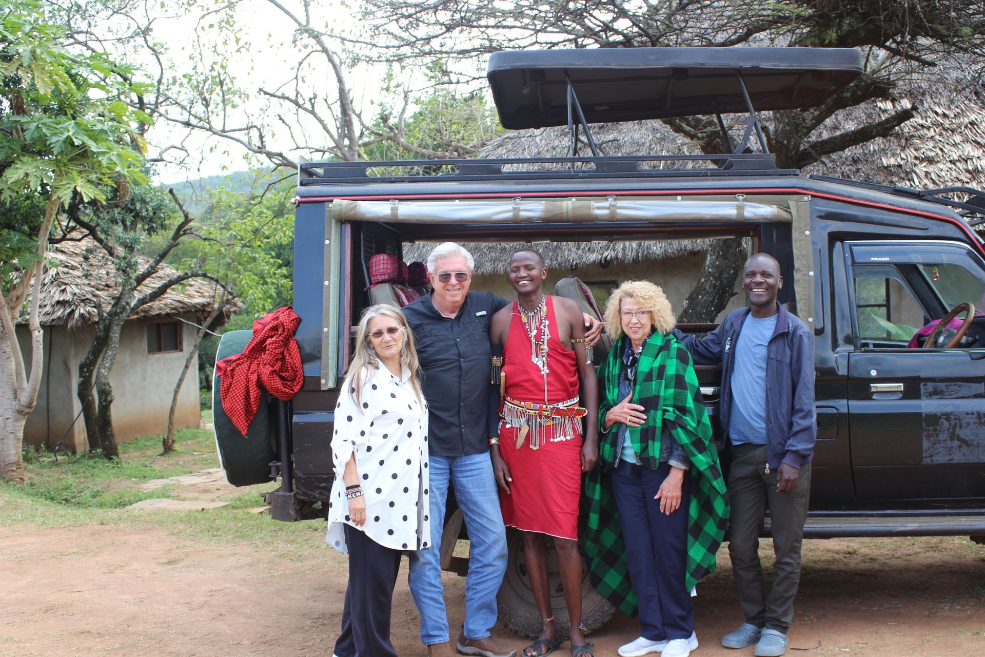 Group of five people posing by a safari vehicle in a village setting. One person wears traditional red Maasai clothing.