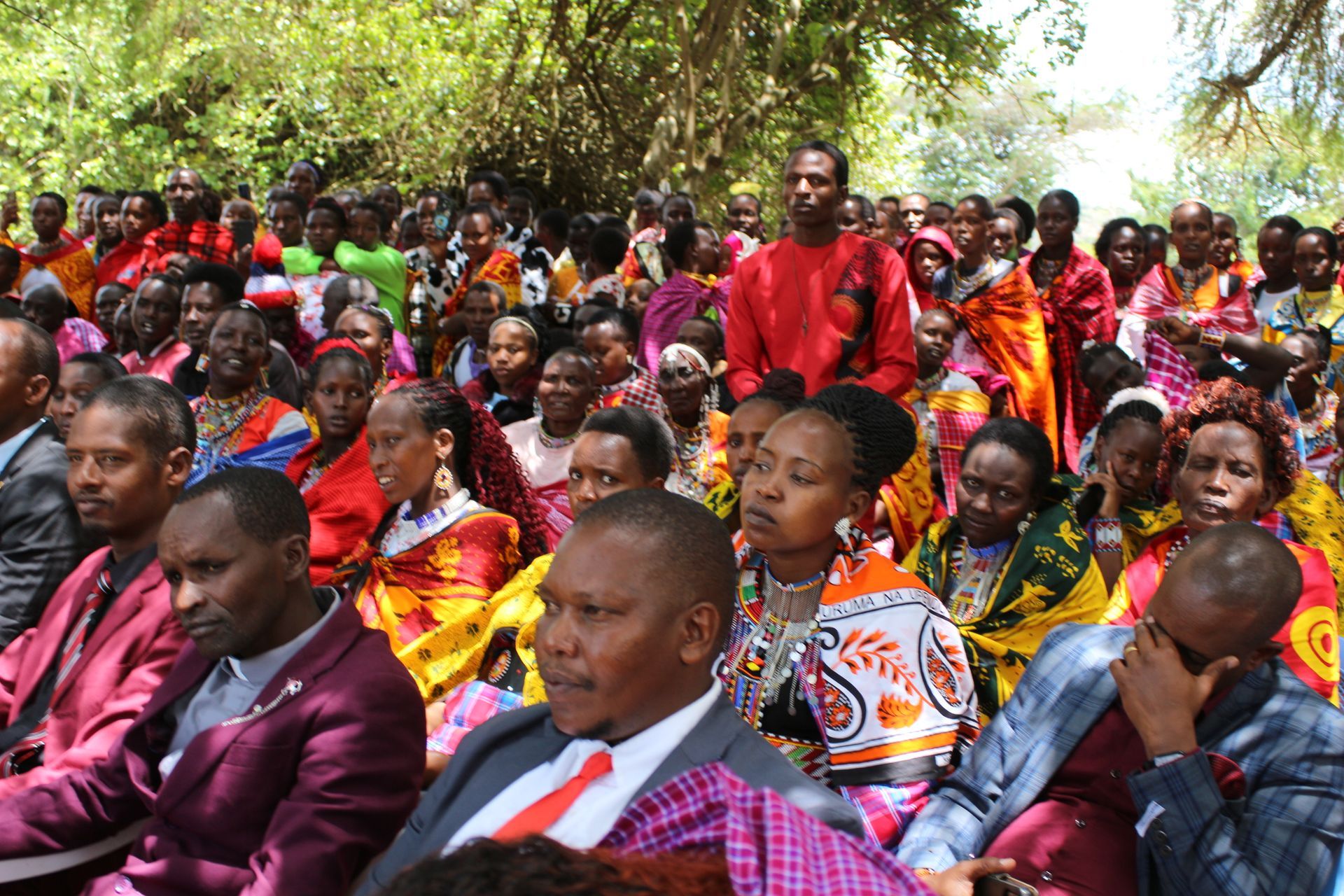Large group of people gathered outdoors, some wearing colorful patterned wraps.