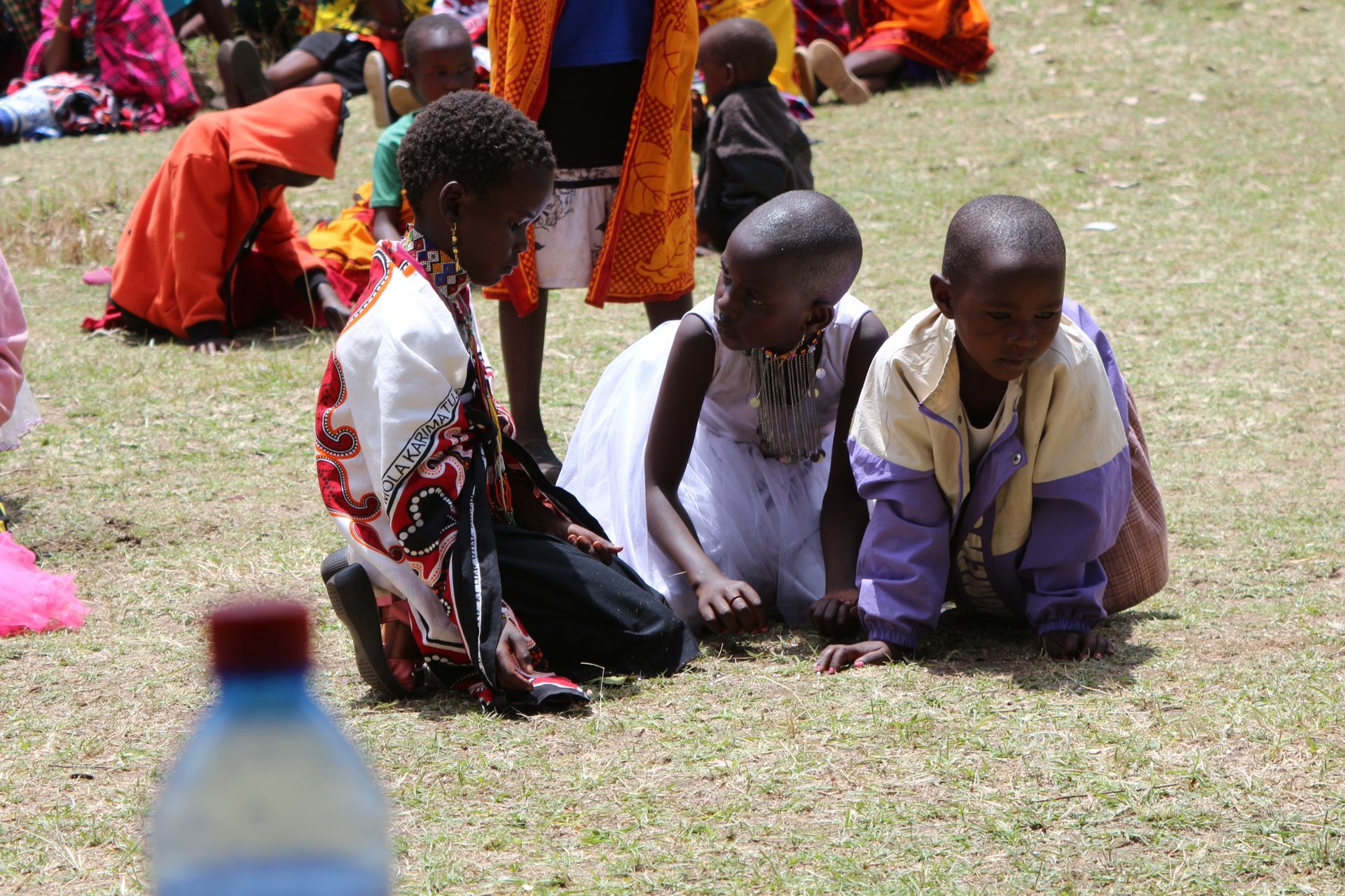Three children kneeling on grass, looking down. Others in colorful clothing in the background.