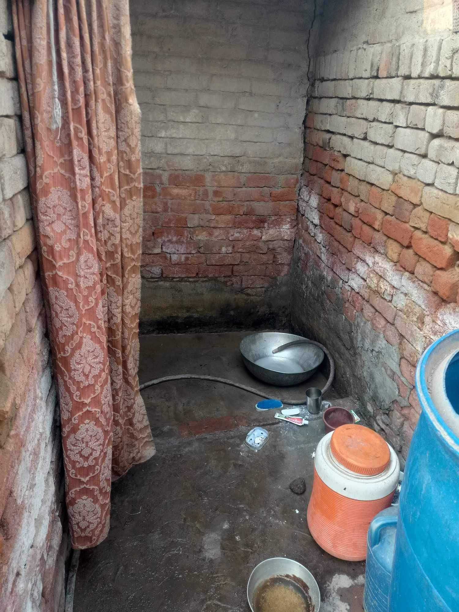 Small, brick-walled outdoor shower with a curtain, metal basin, water jug, and a bucket.