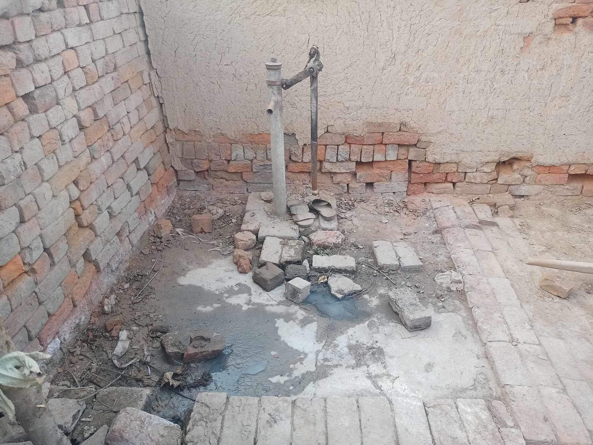 A brick-lined, dirt-floored enclosure with a standing water tap surrounded by broken bricks and mortar.