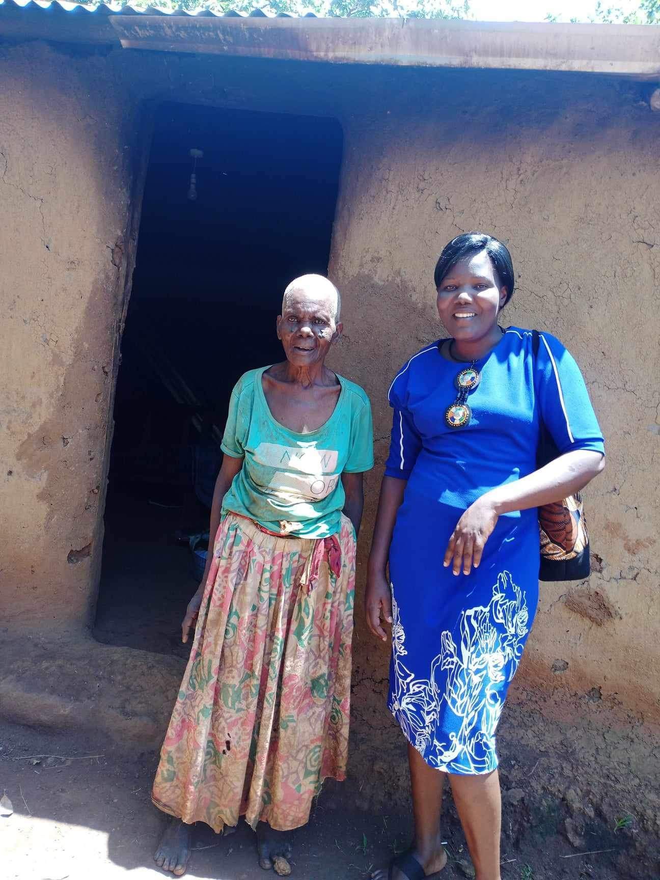 Two women stand in front of a mud hut. The woman on the right wears a blue dress, the other a green shirt and floral skirt.