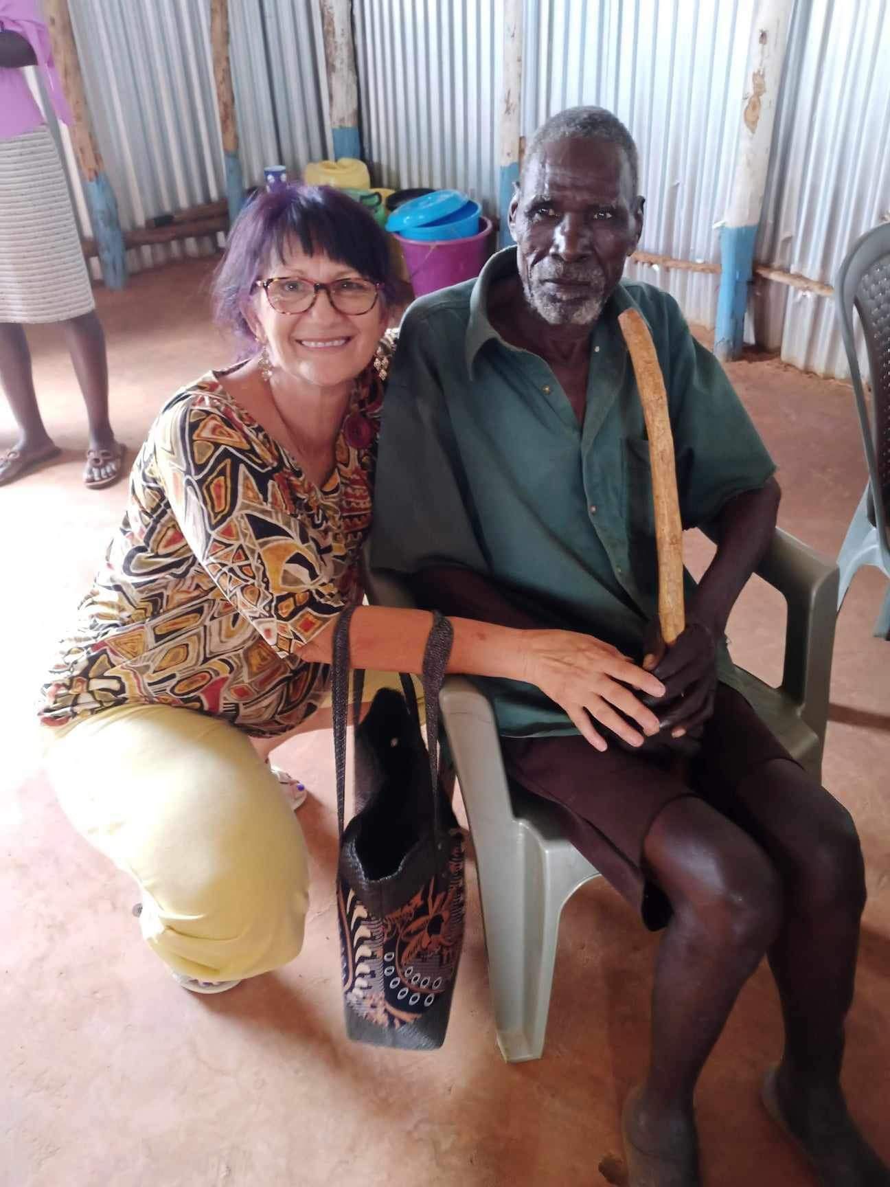 Woman kneels beside a seated person holding a cane. They are indoors. The woman smiles, wearing glasses and patterned top.