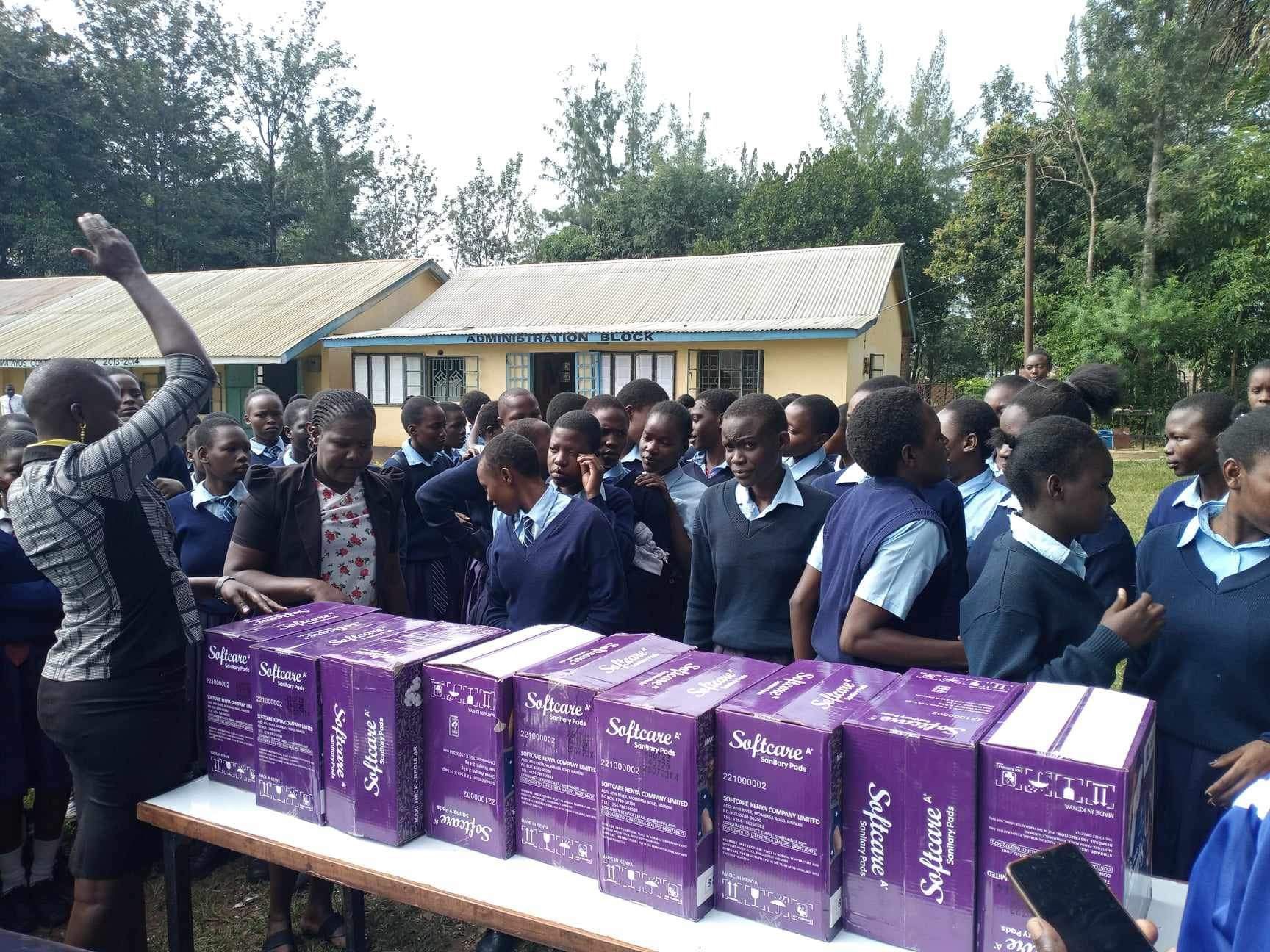 People gathered outdoors near a building with boxes of purple supplies. A person gestures.