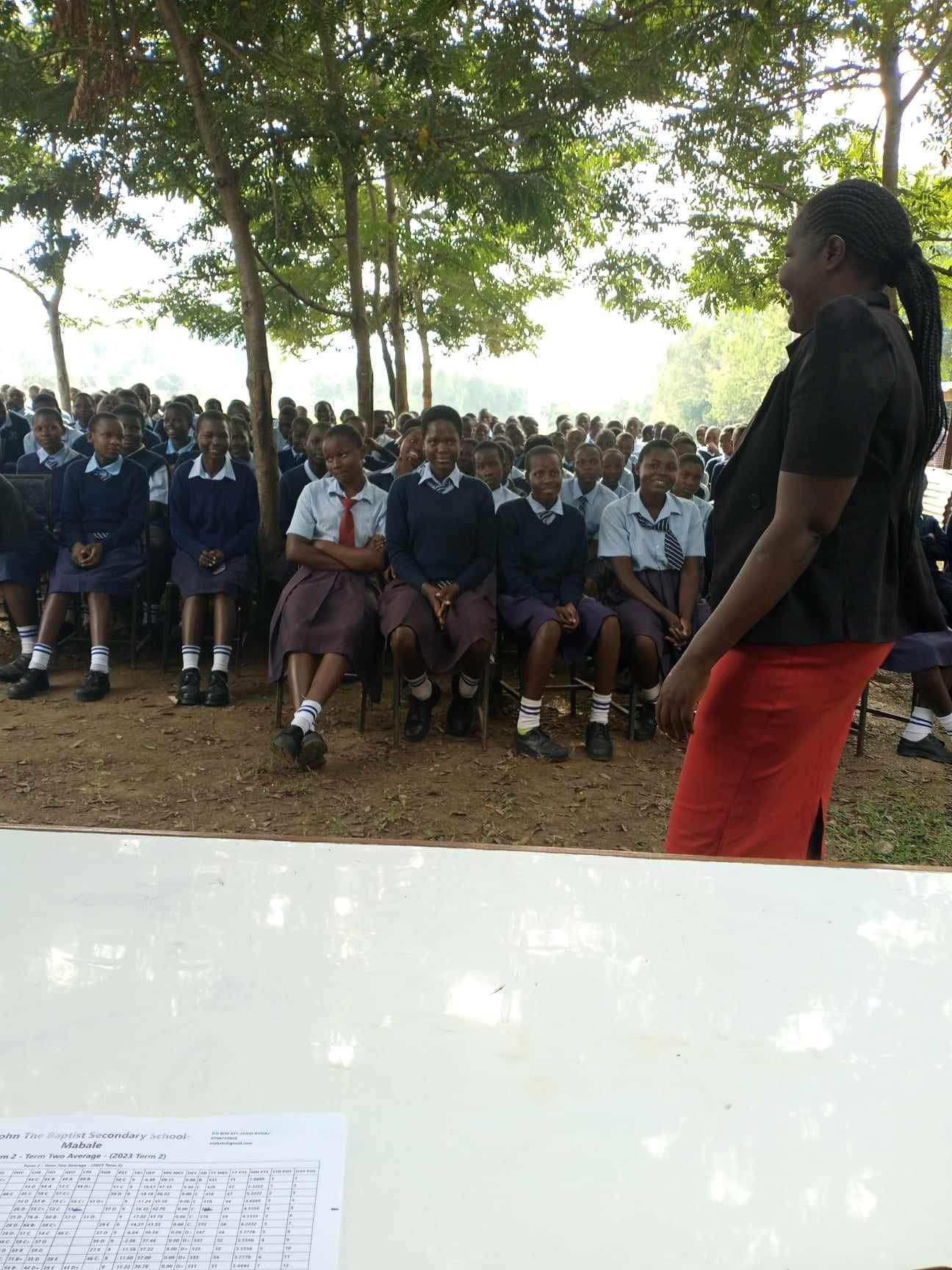 Woman addressing a group of students seated outdoors. Students wear school uniforms.