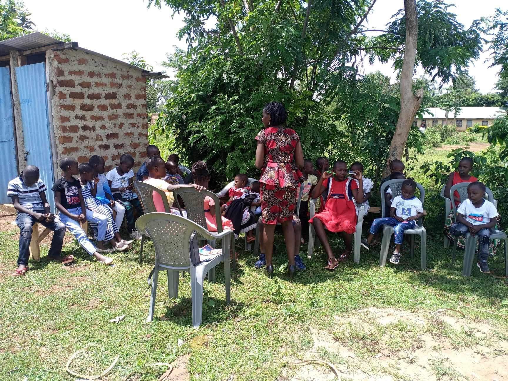 A woman speaks to a group of children seated in chairs outside a building, under trees.