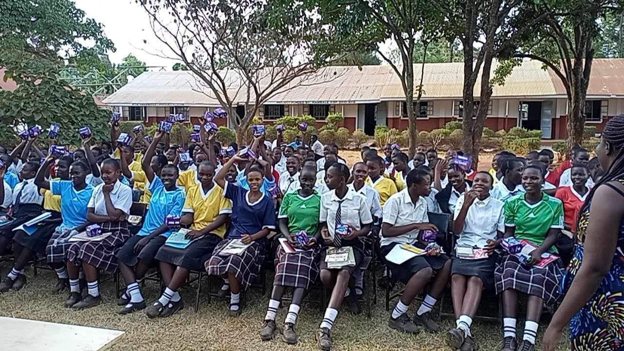 Schoolchildren seated outdoors, many raising hands, buildings in background.