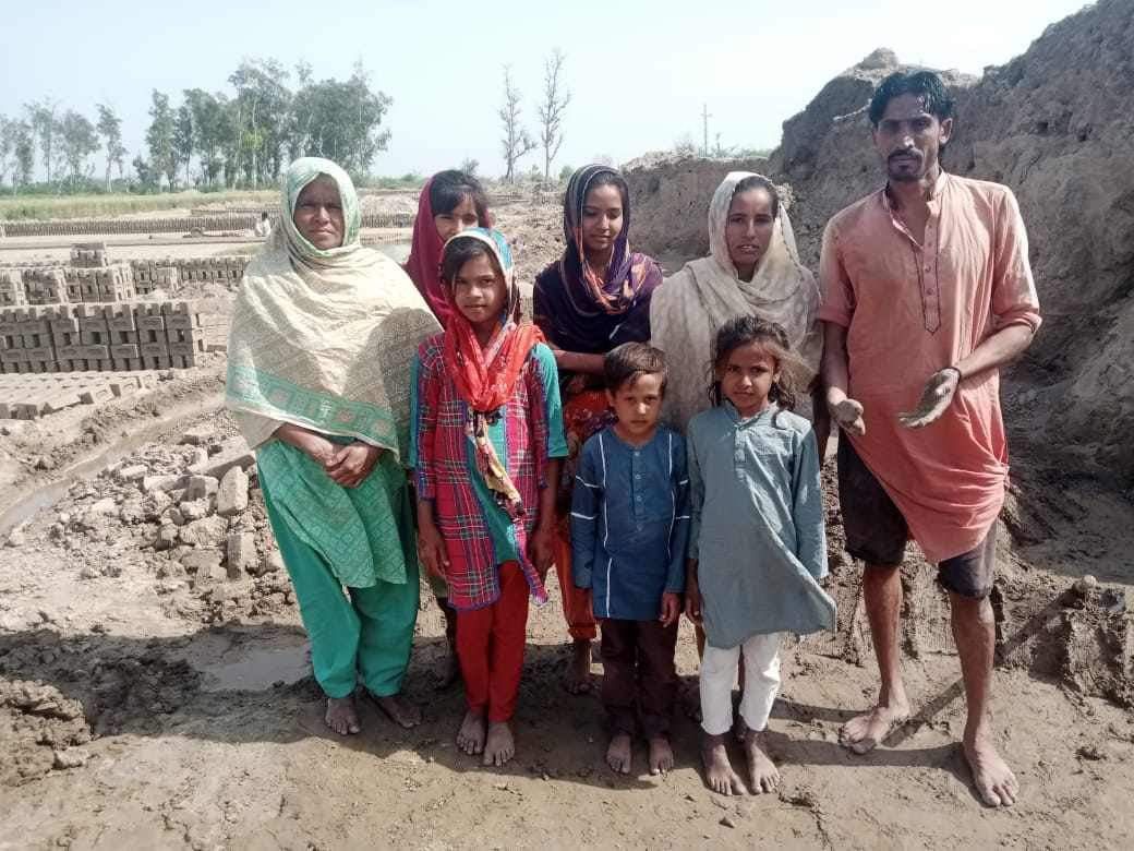Family of seven standing in a brickyard. Several women wearing headscarves, children, and a man posing outdoors.