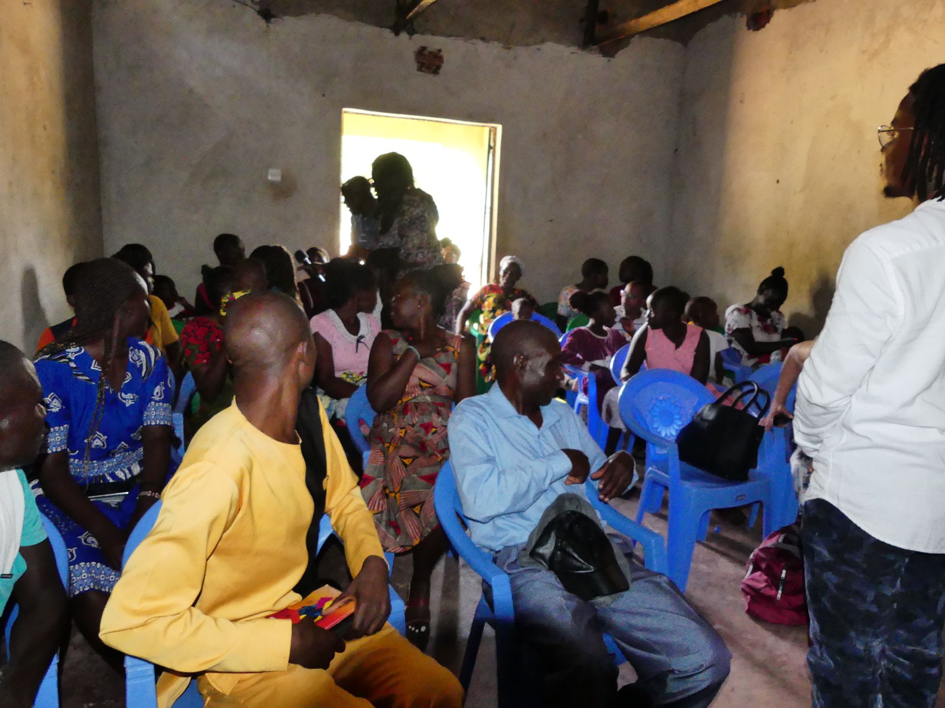 People seated in a room, listening to a presentation. Several blue chairs.