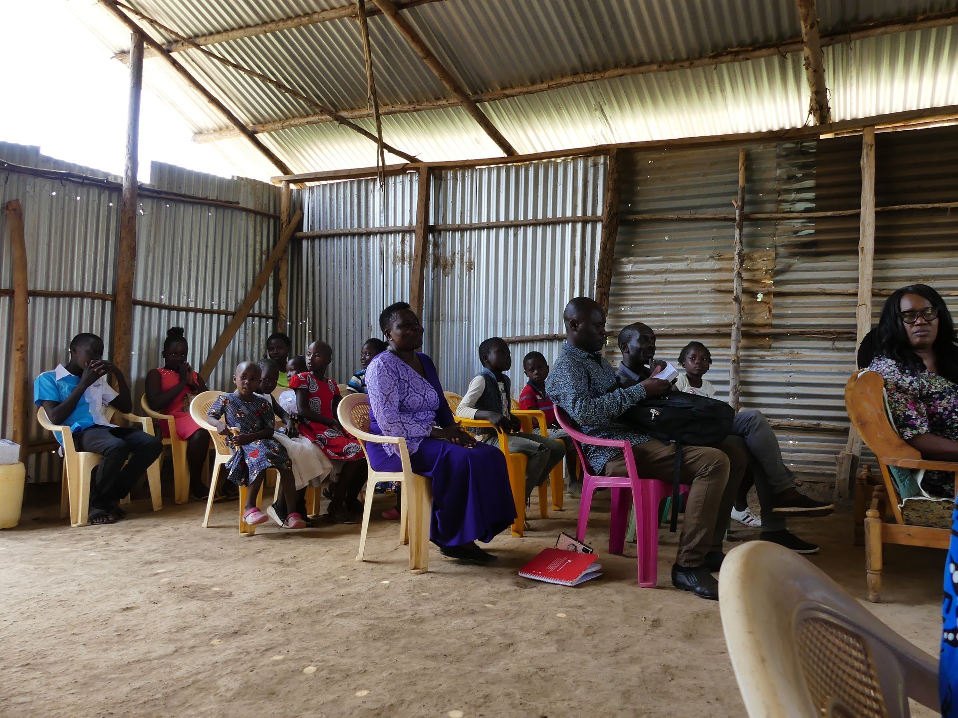 People sitting on chairs inside a simple shelter with corrugated metal walls.