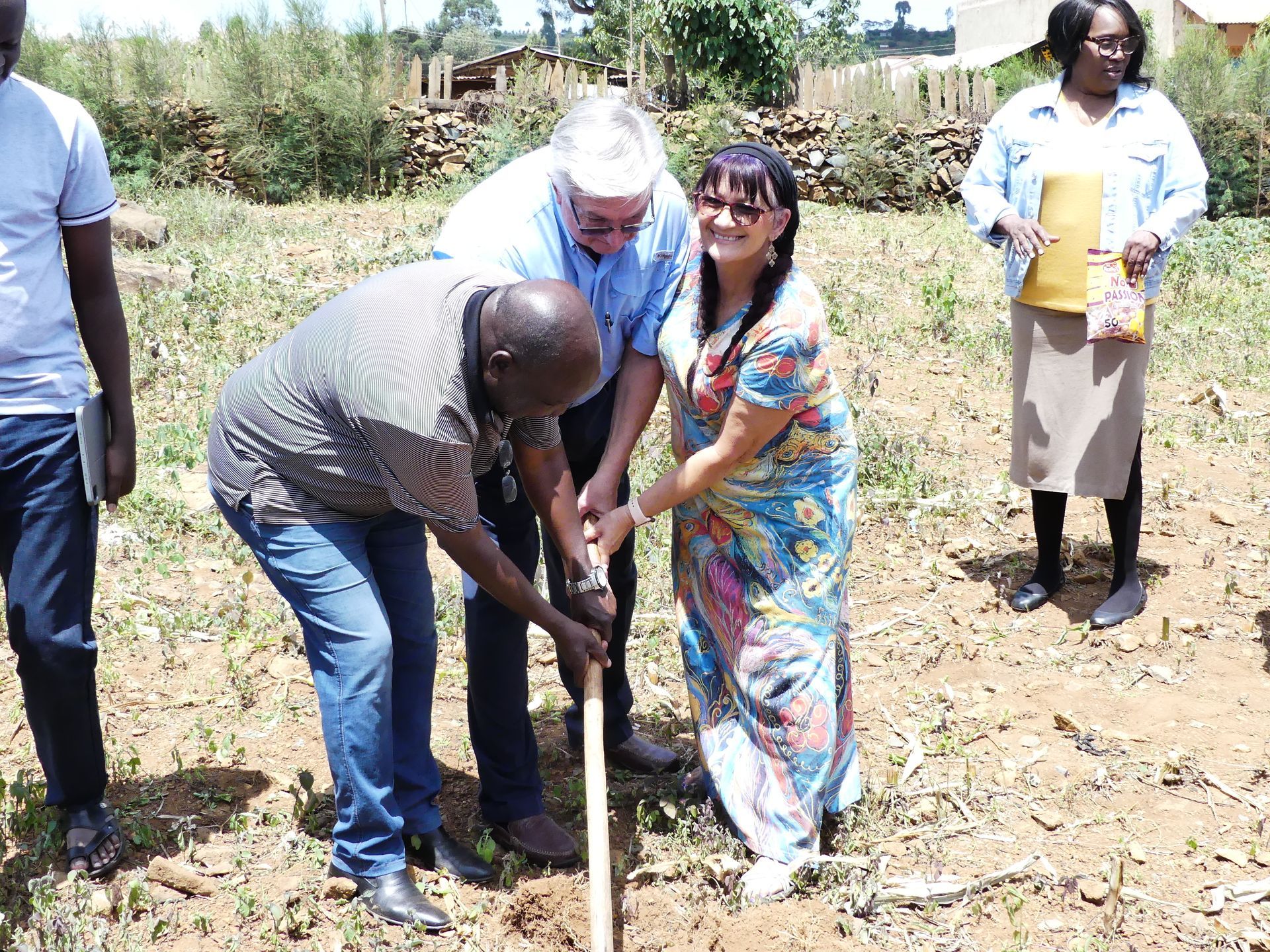 Three people shoveling dirt together outdoors; another woman and man watch.