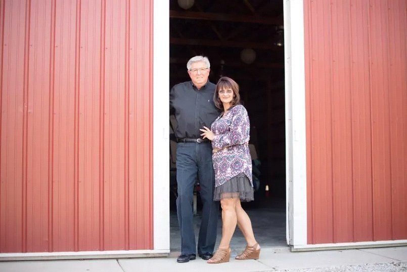 Couple standing in a red barn doorway. Woman in dress and heels, man in black shirt and pants.