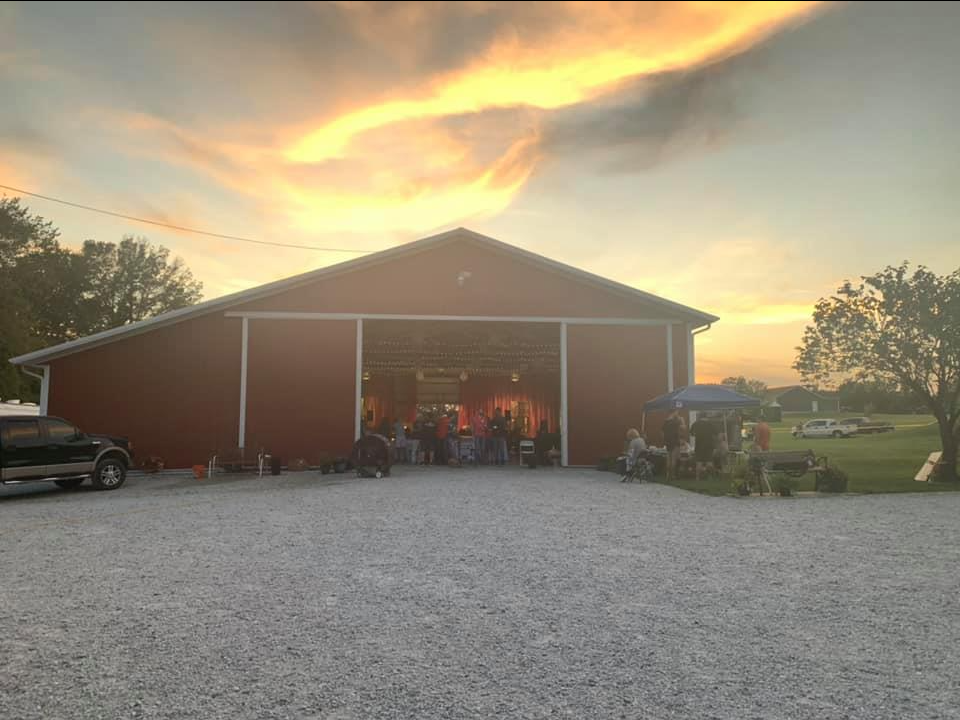 Red barn with open doors, people gathered inside and outside, sunset sky.