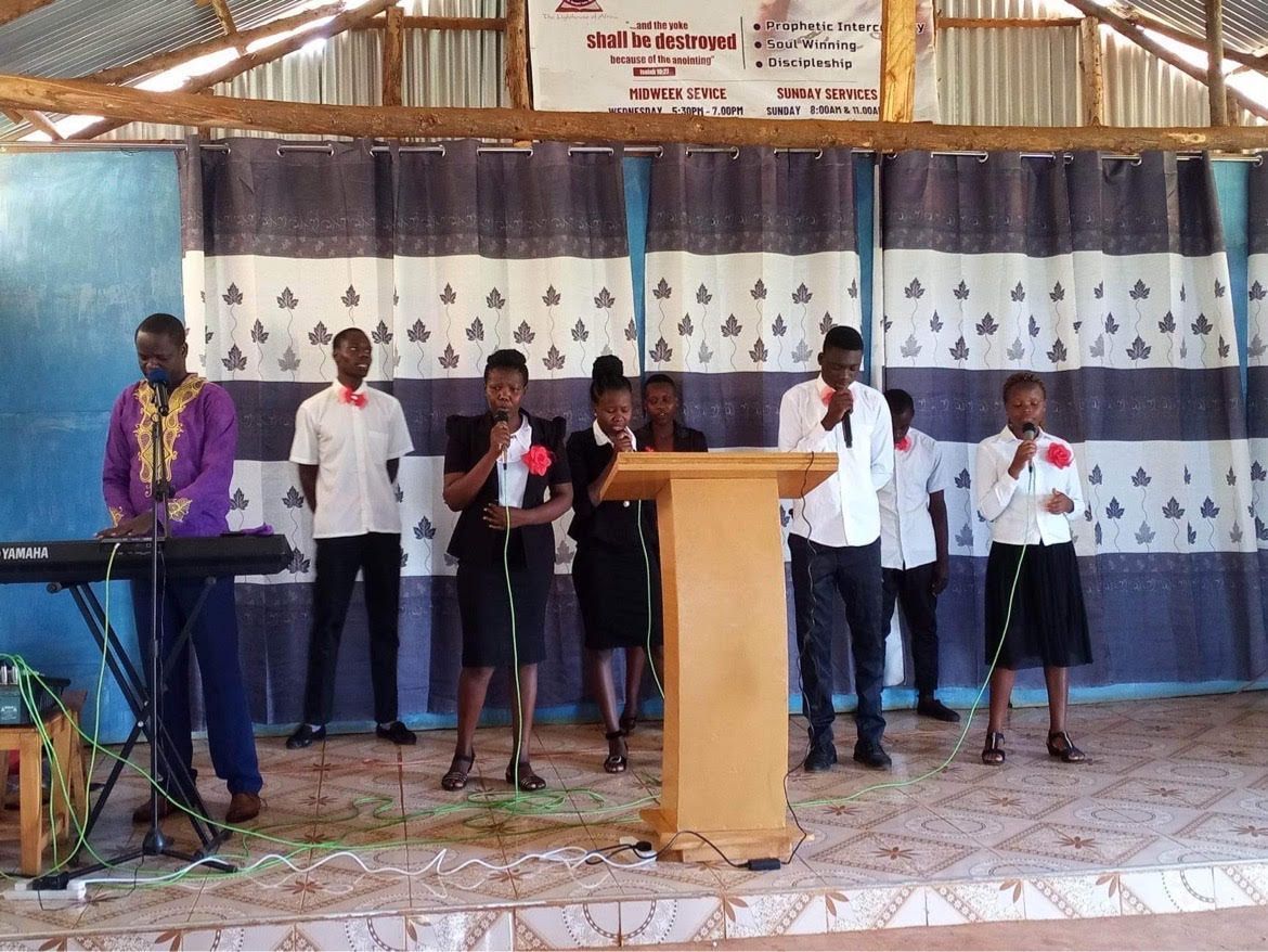 Choir performing on stage. Keyboardist, speakers, and singers with microphones in front of a blue and white backdrop.