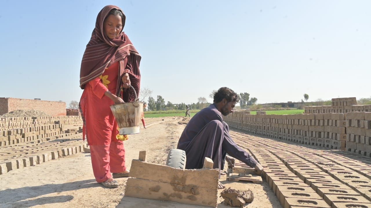Woman carrying bucket, man arranging bricks at an outdoor brickyard. Bright daylight.