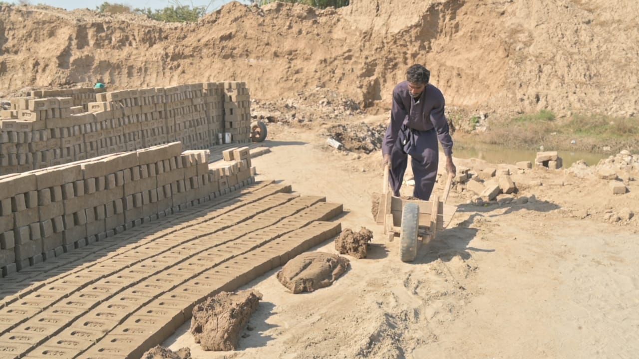 Man pushing a wheelbarrow of bricks in a brickyard. Piles of bricks and earthen embankment in background.