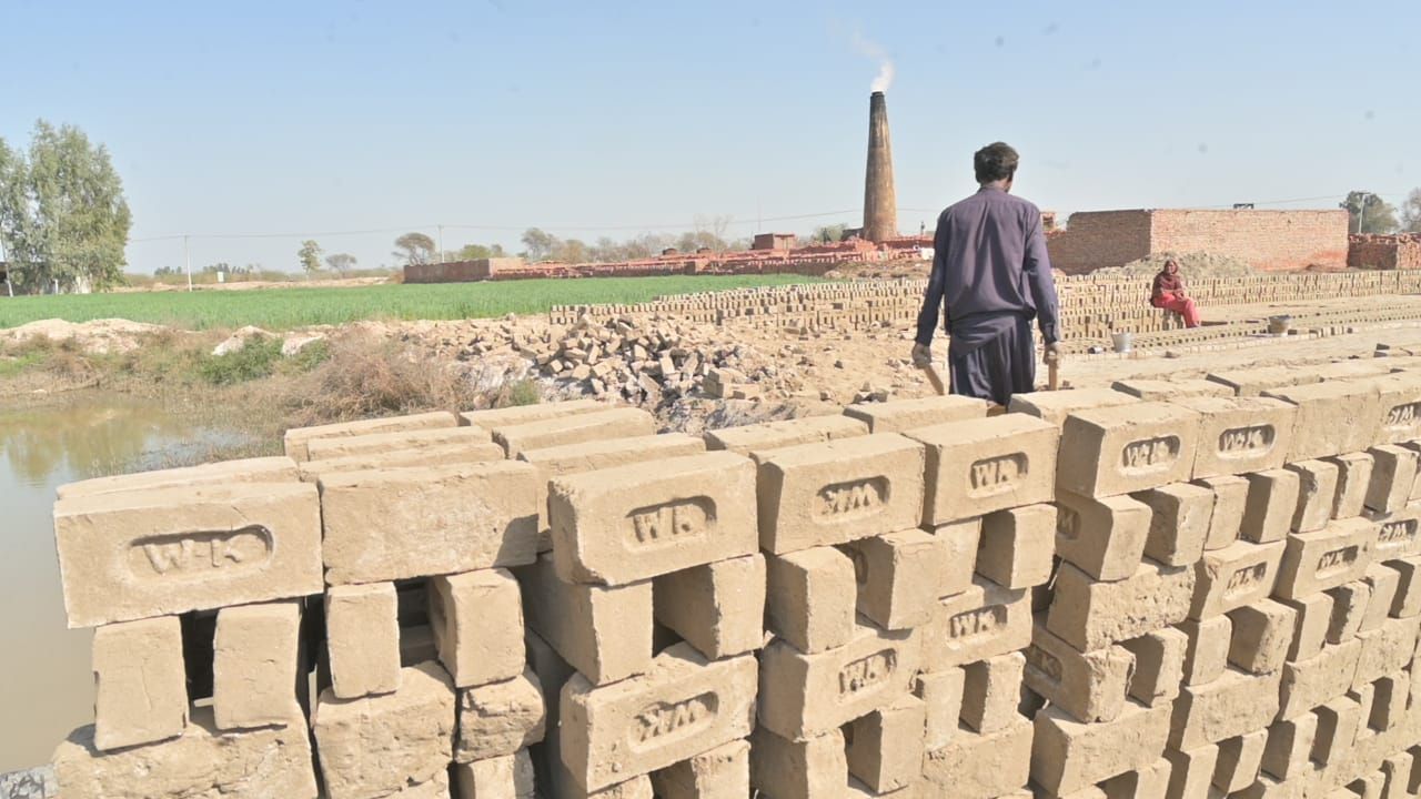 Man walks on bricks stacked near a brickyard; a brick chimney emits smoke.