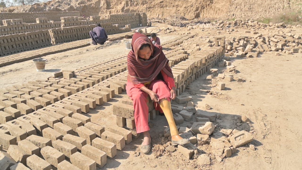 Woman sitting on brick pile, working at a brickyard, surrounded by rows of bricks and a worker in the background.