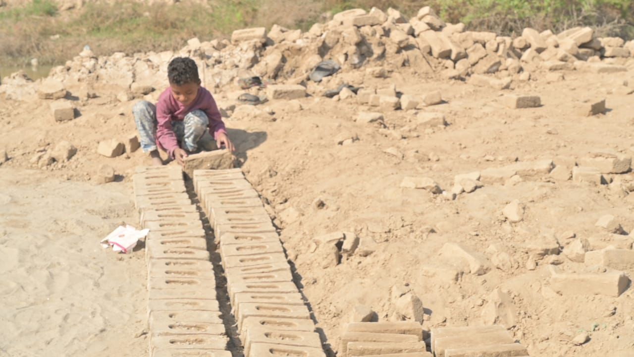 Child arranging bricks on a sandy ground; other bricks and dirt piles in background.